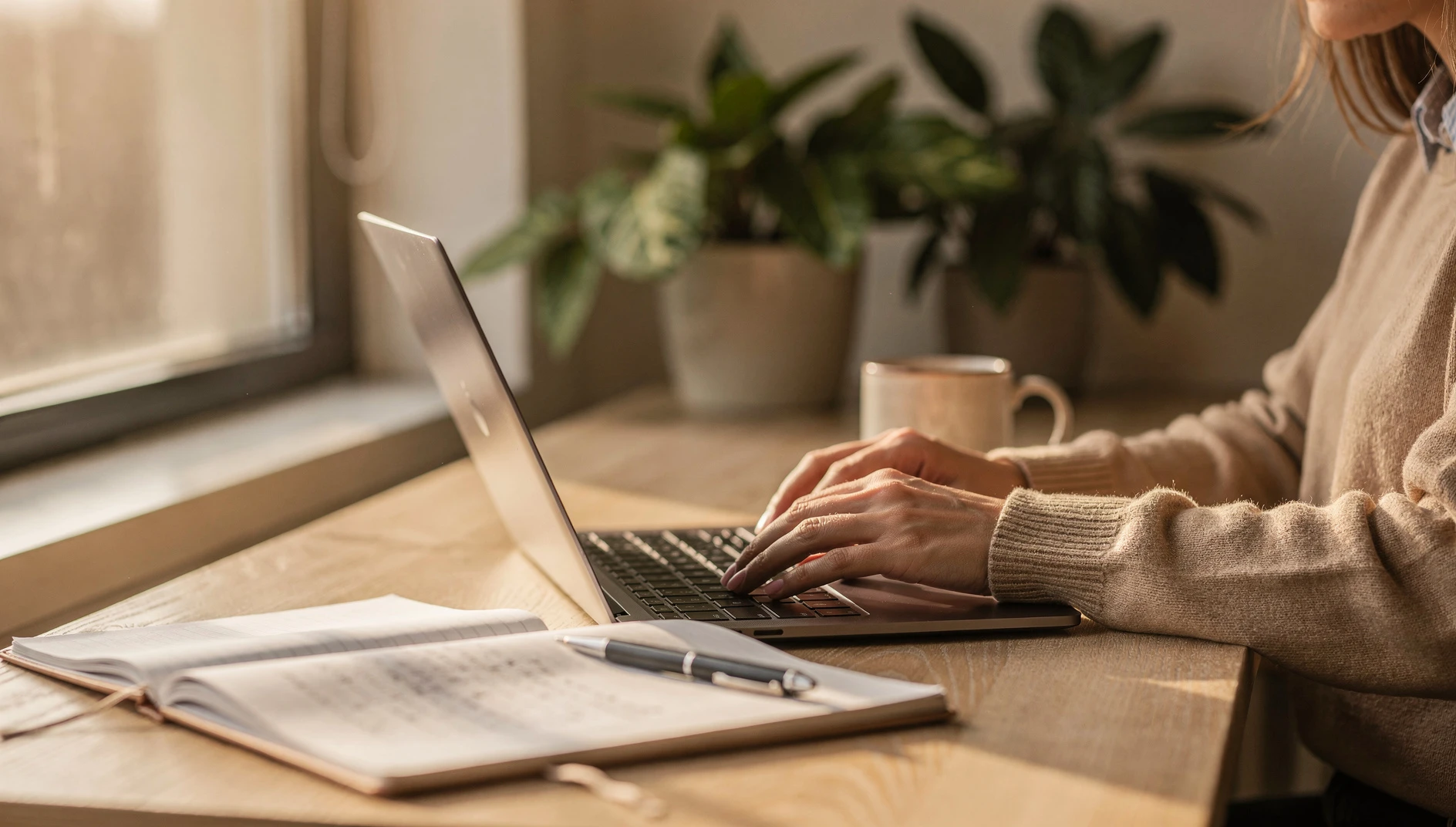 A person writing at a light wooden desk with a notebook open beside them in warm morning light