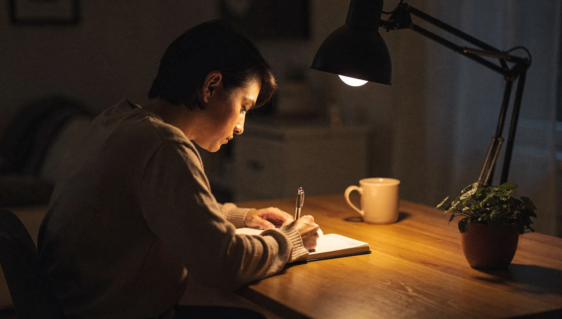 A person writing in a journal at a home desk in warm lamp light in the evening