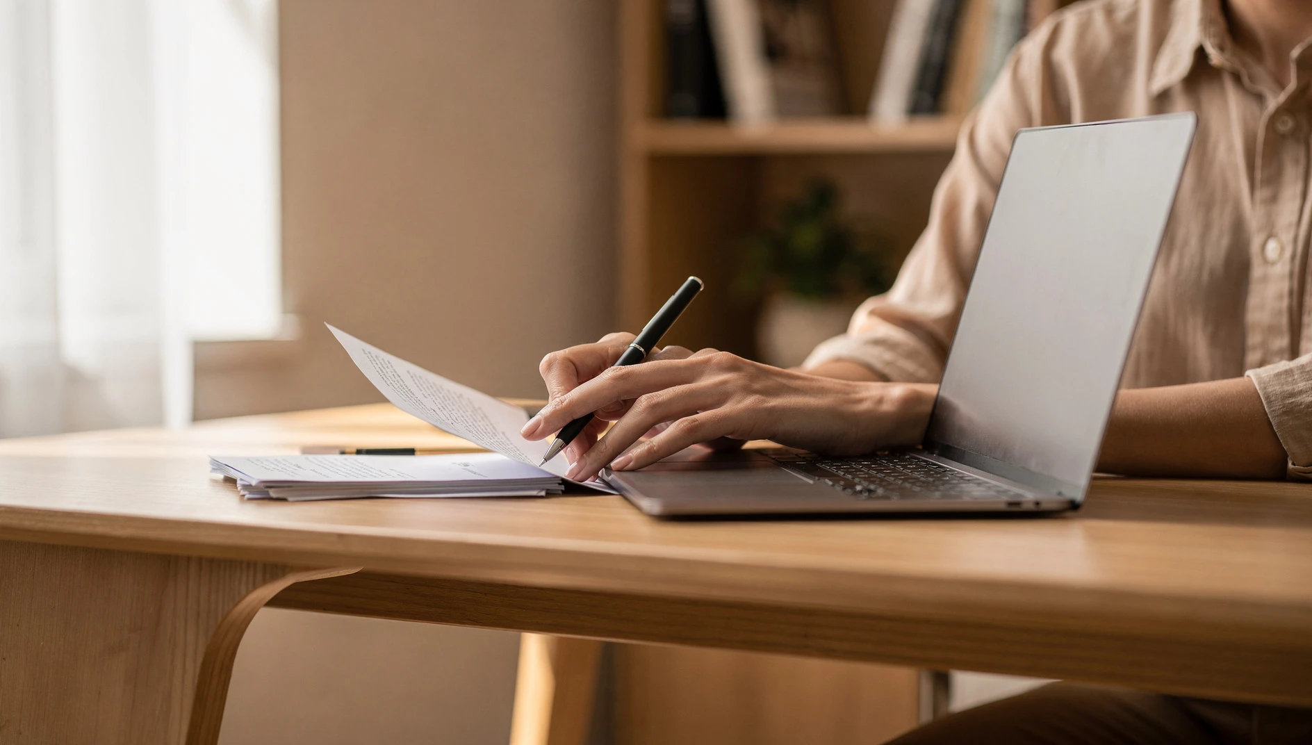 A professional reviewing business documents at a desk with a laptop and natural light from a window