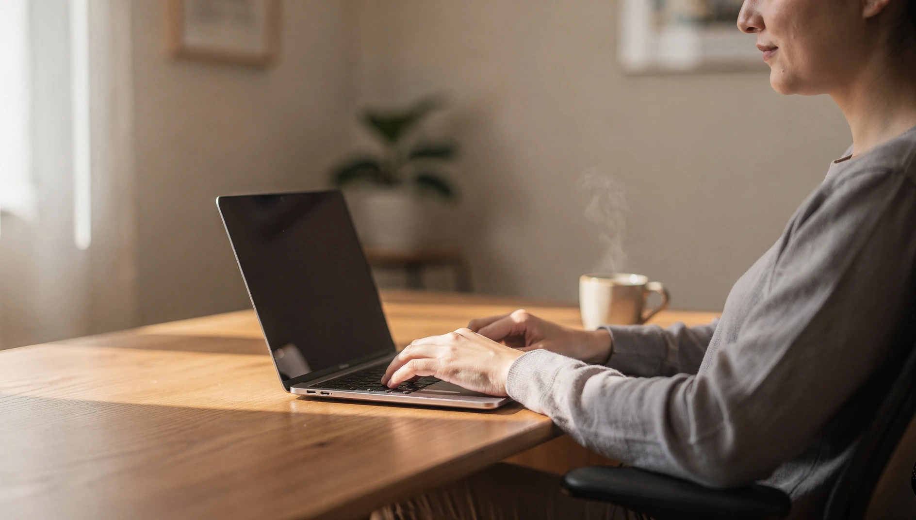 A person typing on a laptop at a clean wooden desk with a coffee cup in soft window light