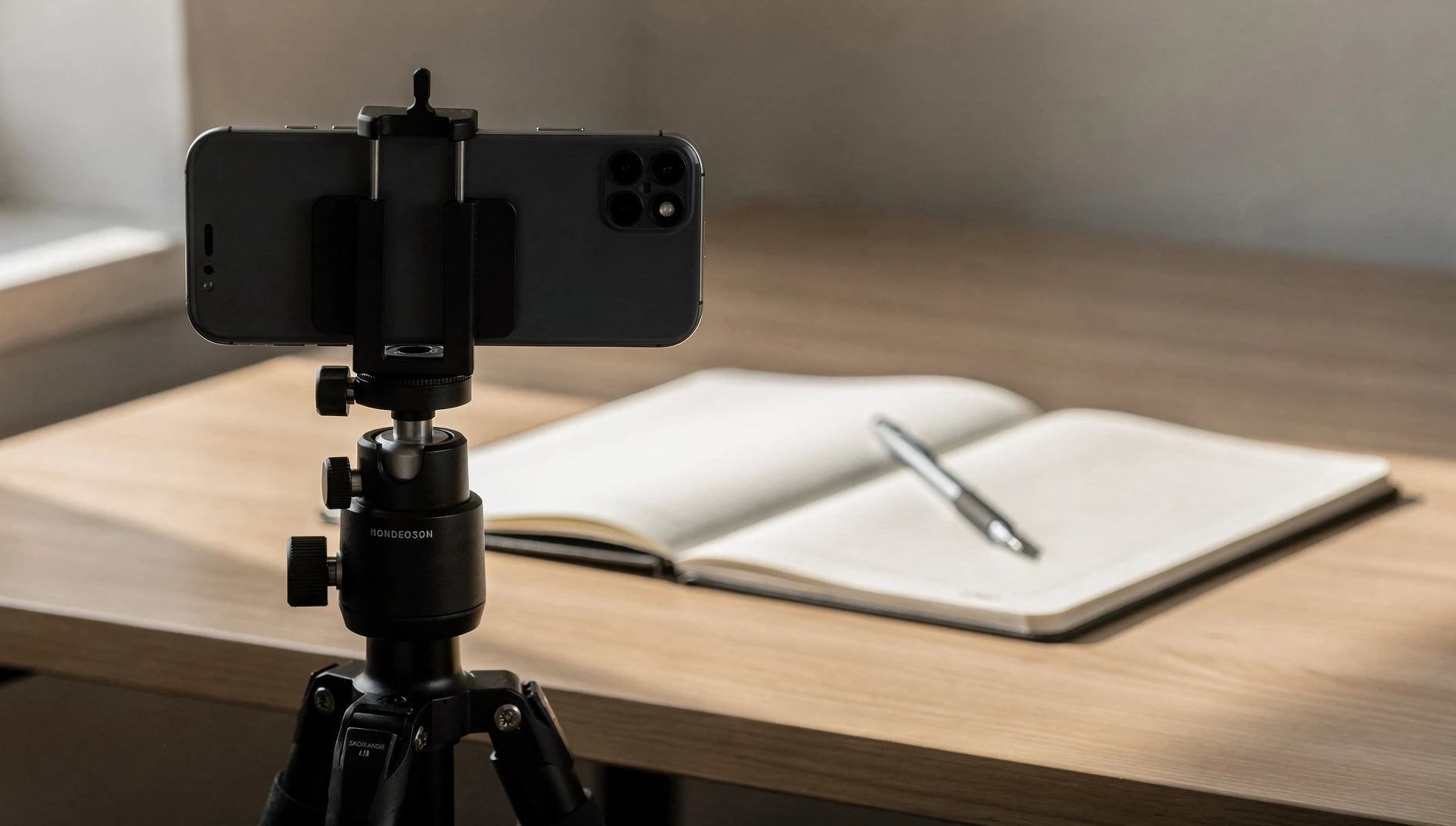A smartphone on a tripod pointed at a desk with an open notebook and pen in soft warm window light, no person visible