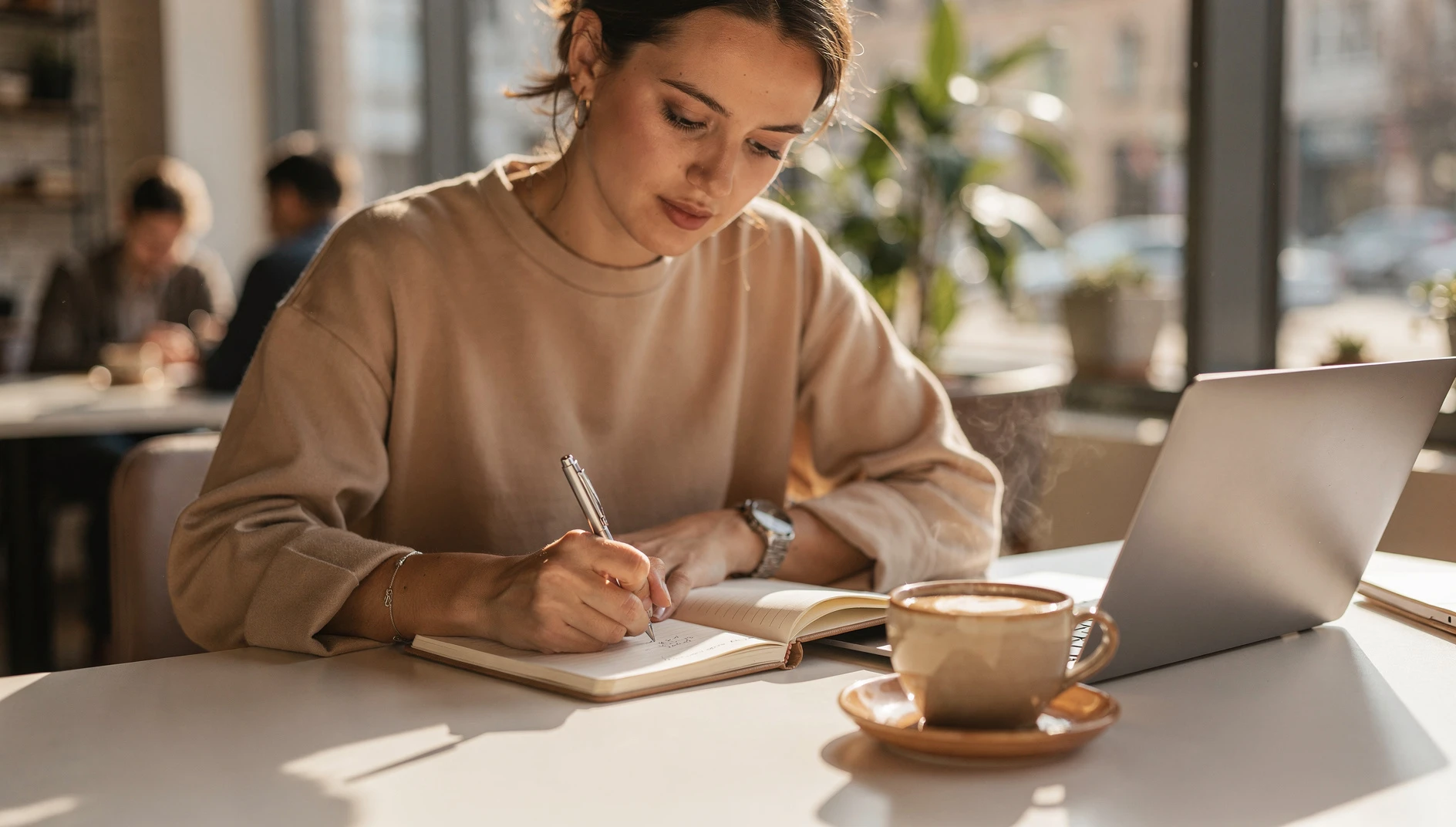 A person writing in a notebook at a bright cafe table with a laptop and coffee in warm morning light