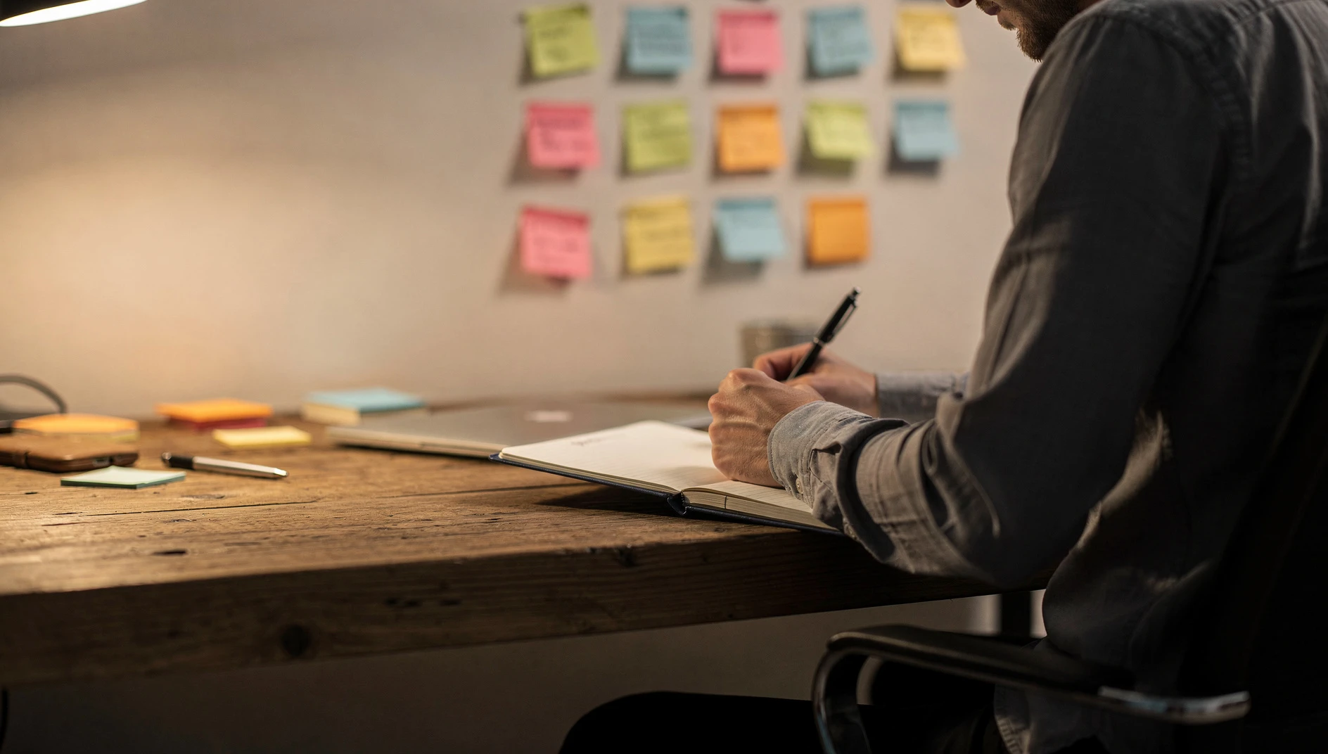 A person writing in a notebook at a wooden desk with sticky notes on a wall during a brainstorming session