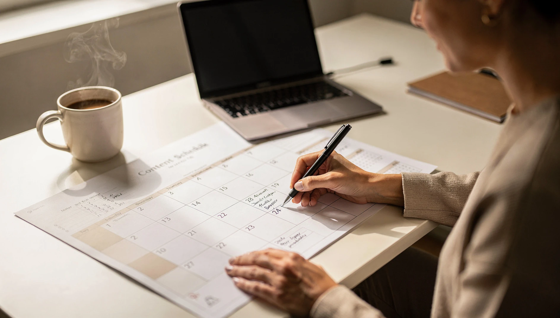 A person planning a content calendar on a physical calendar at a bright desk with coffee and laptop nearby