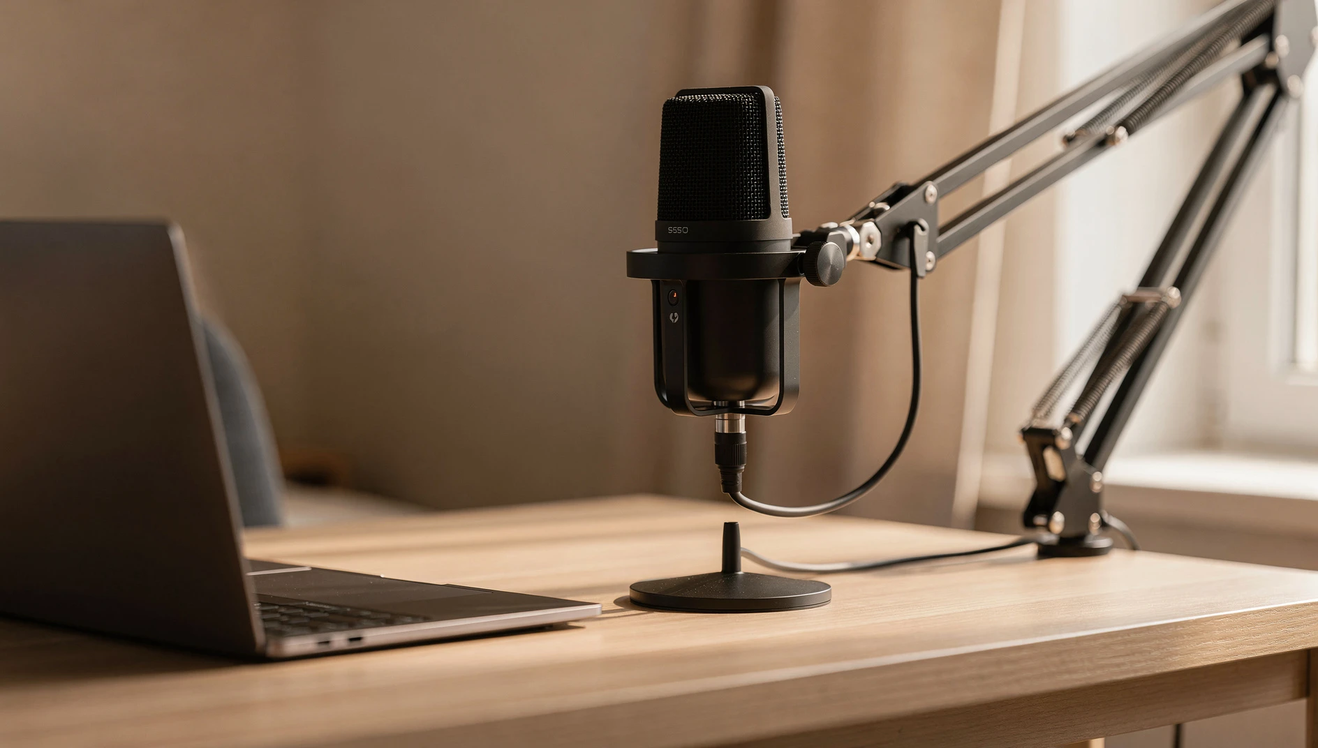 A podcast microphone on a desk arm beside a laptop in a minimal home office with soft natural window light