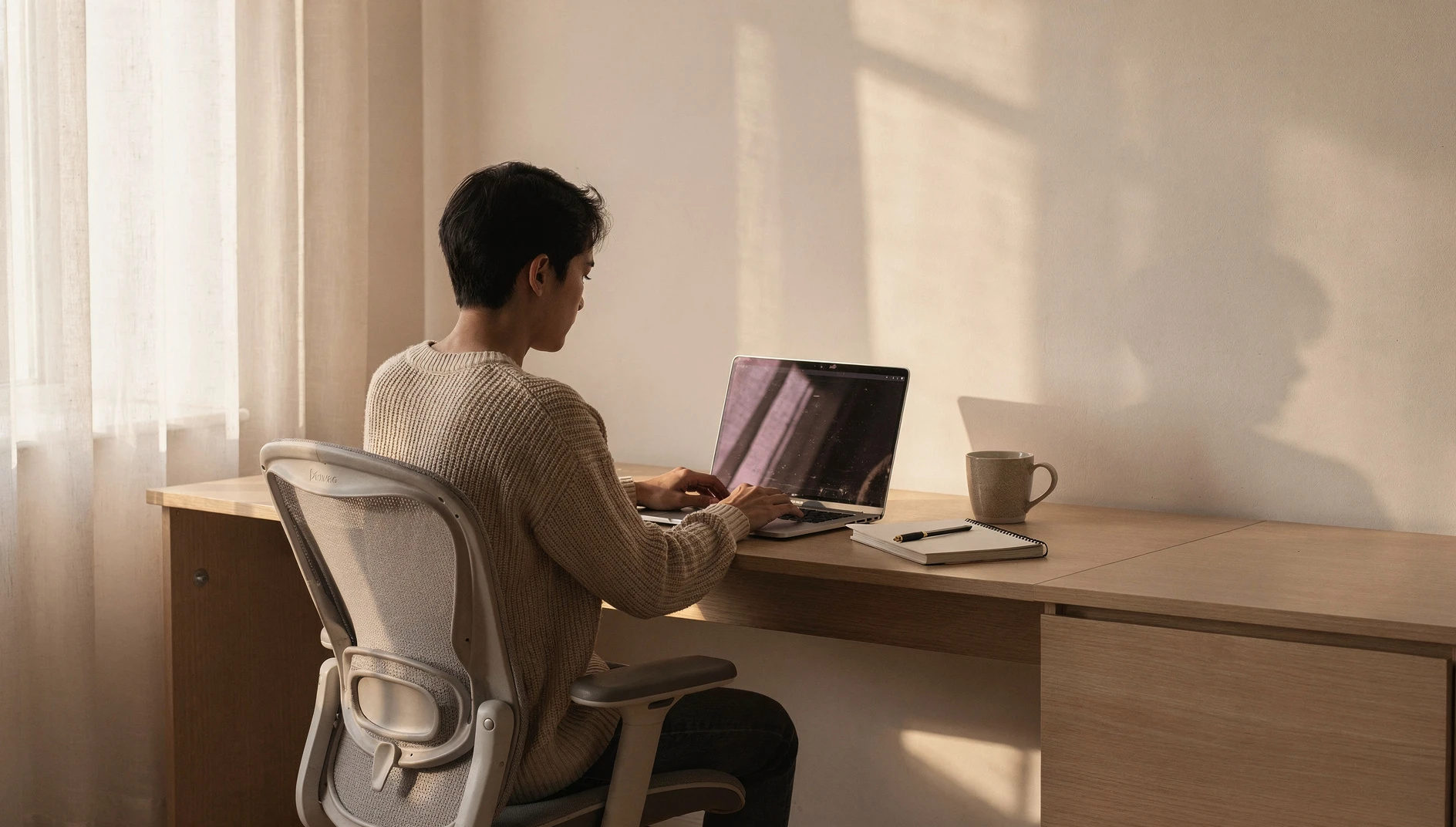 A professional working on a laptop at a home desk near a window with morning natural light and a minimalist setup