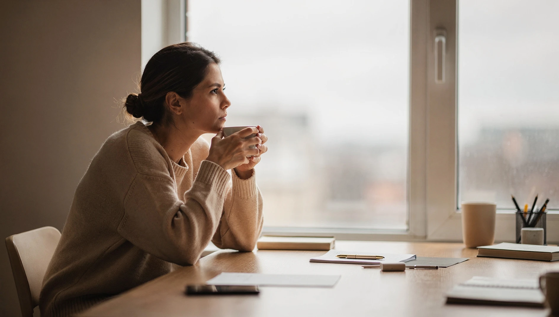 A person looking out a window at a desk with a coffee cup in soft natural light