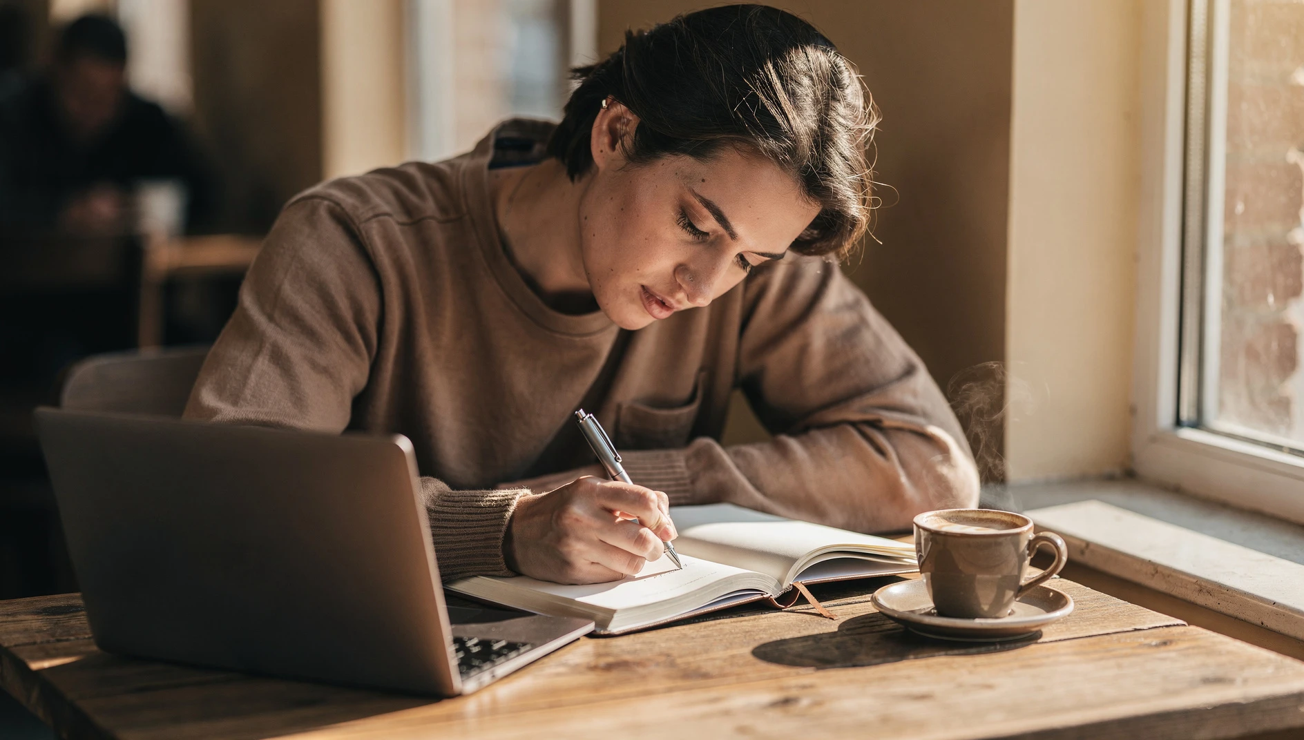 A person writing in a notebook at a cafe table with a laptop and coffee in warm afternoon light